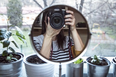 Woman in mirror holding up a camera, with outdoor window in background